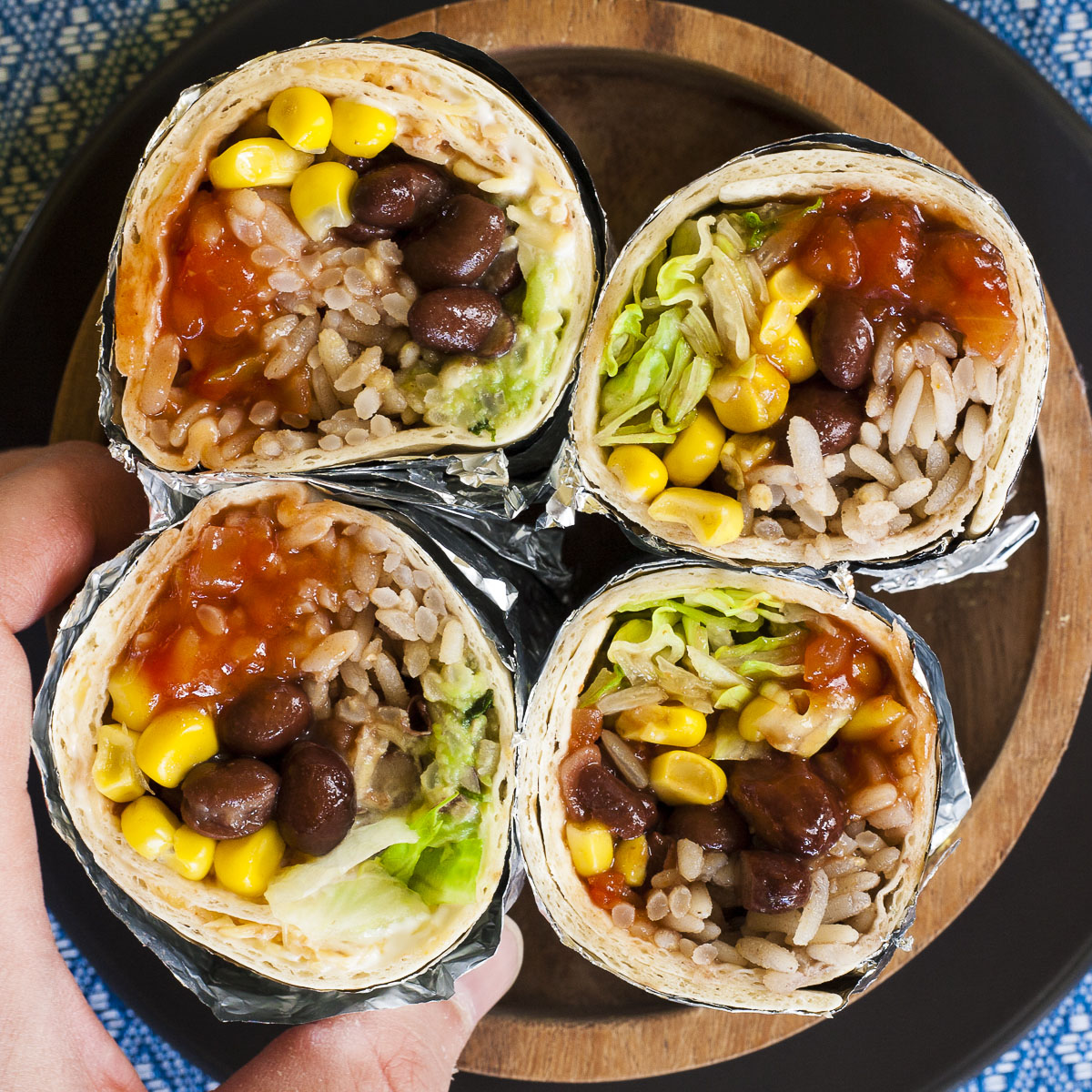 A hand is holding one of the four rainbow coloured round-shaped rolls from above. Inside red, yellow, brown, green and white pieces.