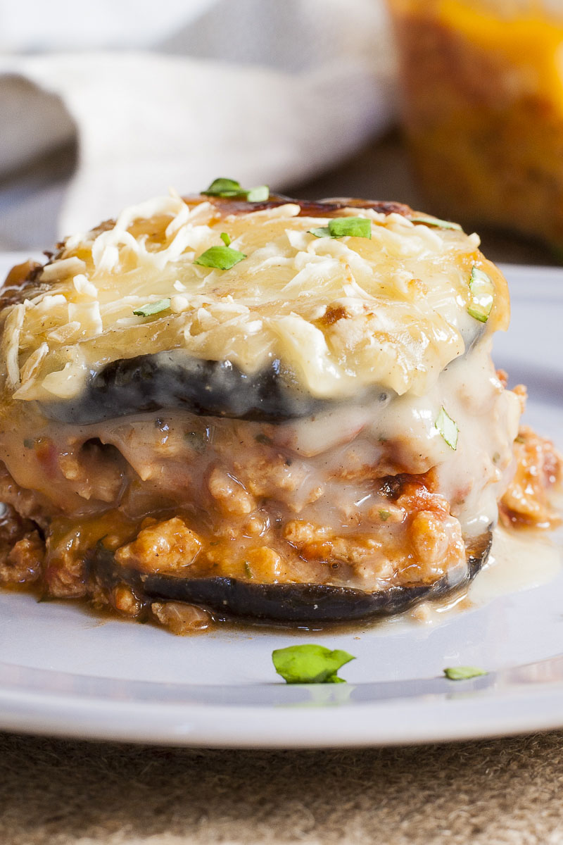 One serving of vegan moussaka is on a light purple plate. You can see the layers of purple eggplant, brown crumbles and white sauce on top. 