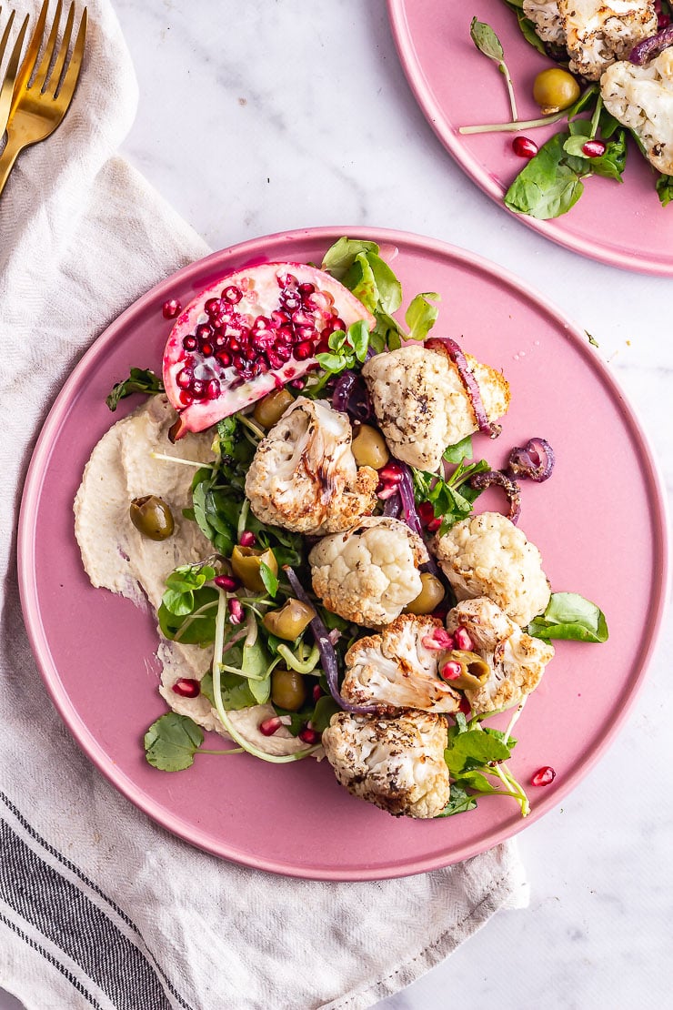 Vibrant pink plate from above with roasted cauliflower florets, olives, fresh green leaves, purple onion slices, pomegranate seeds and hummus on the bottom. 