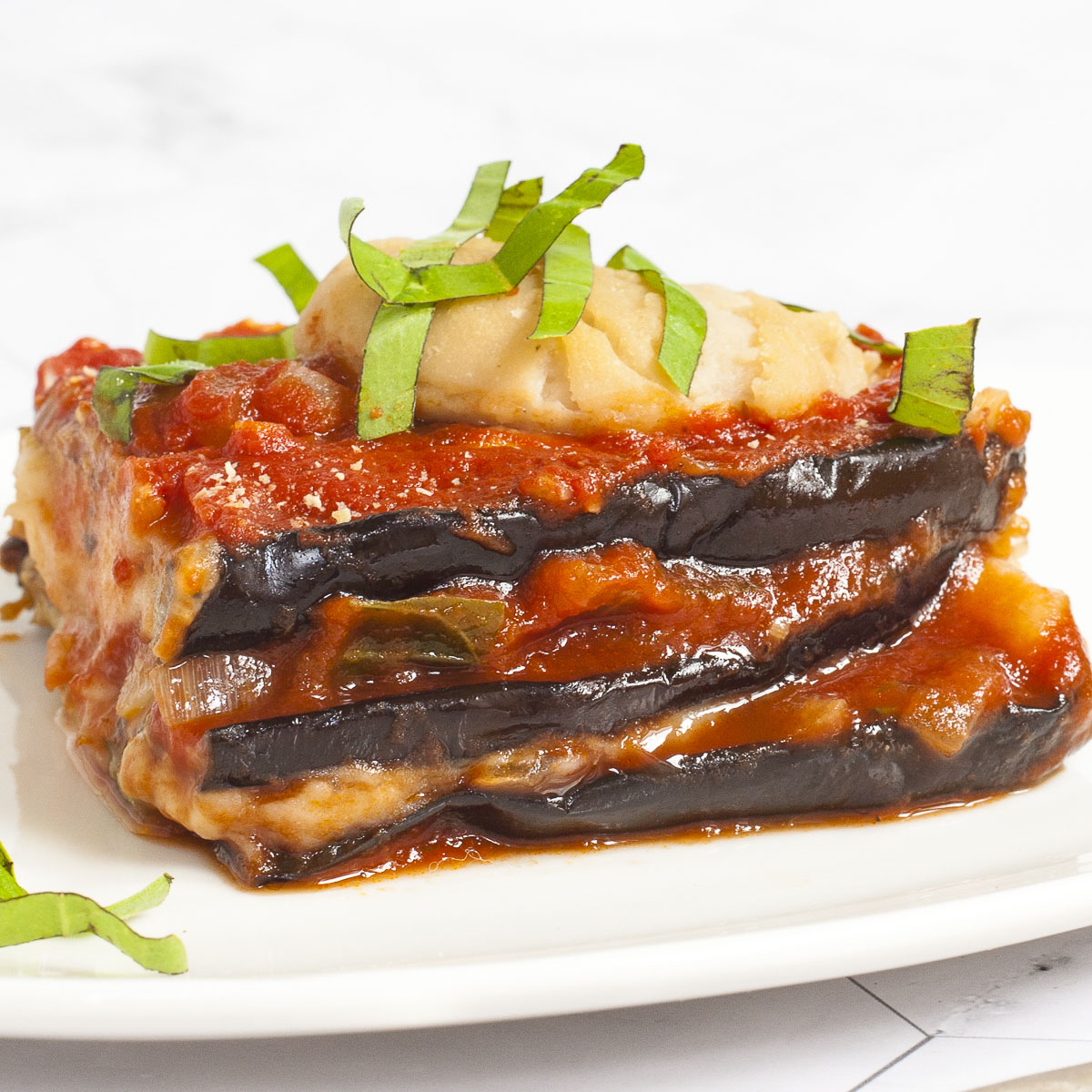 3 slices of purple eggplant in between red and white sauces on a white plate topped with fresh green herbs. In the background there is a red casserole