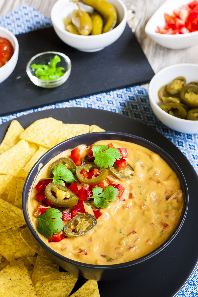 Yellow sauce in a black bowl topped with red veggie chunks, sliced green pepper and green leaves of herbs. Several small white bowls behind with different red and green toppings