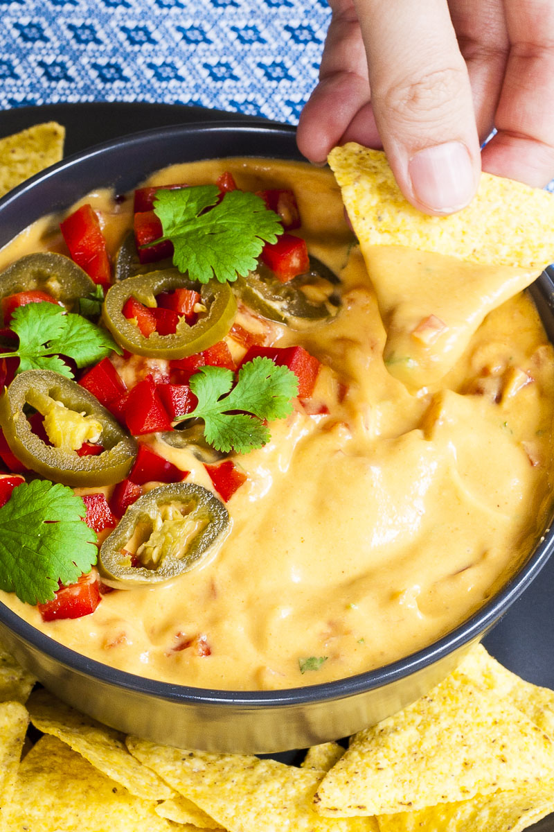 Yellow sauce in a black bowl topped with red veggie chunks, sliced green pepper and green leaves of herbs. A tortilla chips is dipped in it. Other tortilla chips are scattered around.