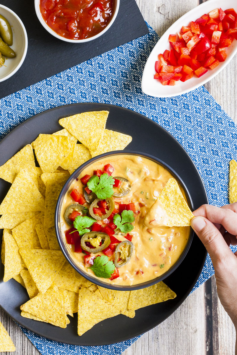 Yellow sauce in a black bowl topped with red veggie chunks, sliced green pepper and green leaves of herbs. A tortilla chips is dipped in it. Other tortilla chips are scattered around. Several small white bowls behind with different red and green toppings