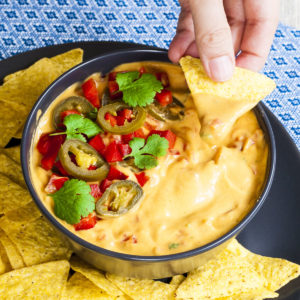 Yellow sauce in a black bowl topped with red veggie chunks, sliced green pepper and green leaves of herbs. A tortilla chips is dipped in it. Other tortilla chips are scattered around.