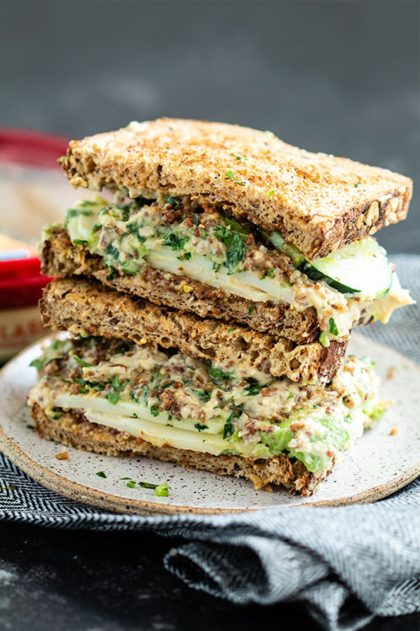 Stack of sandwich on a gray plate with brown seeded bread slices, green veggies and tabbouleh spread. 