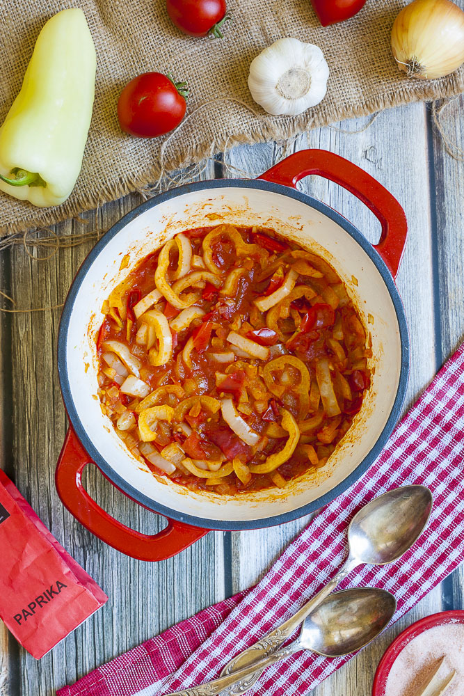 Red and yellow veggie stripes in a red stew in a red and white pot. Ingredients of the lecso scattered around like wax pepper, tomato, onion, garlic and paprika powder in a red paper bag.