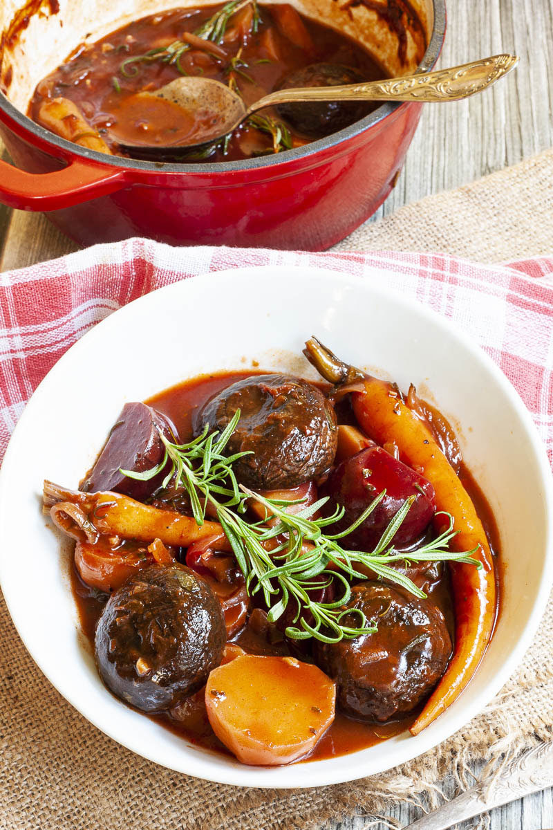 White plate with yellow, red, purple veggies and portobello mushroom caps topped with fresh green herbs on twigs. Red pot is behind it