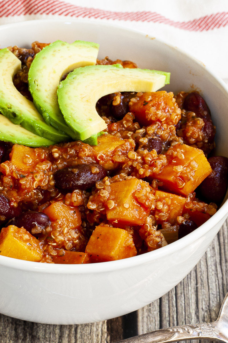 In a small white bowl orange cubes, dark brown beans, small white, yellow seeds with 3 slices of green avocado on top. 