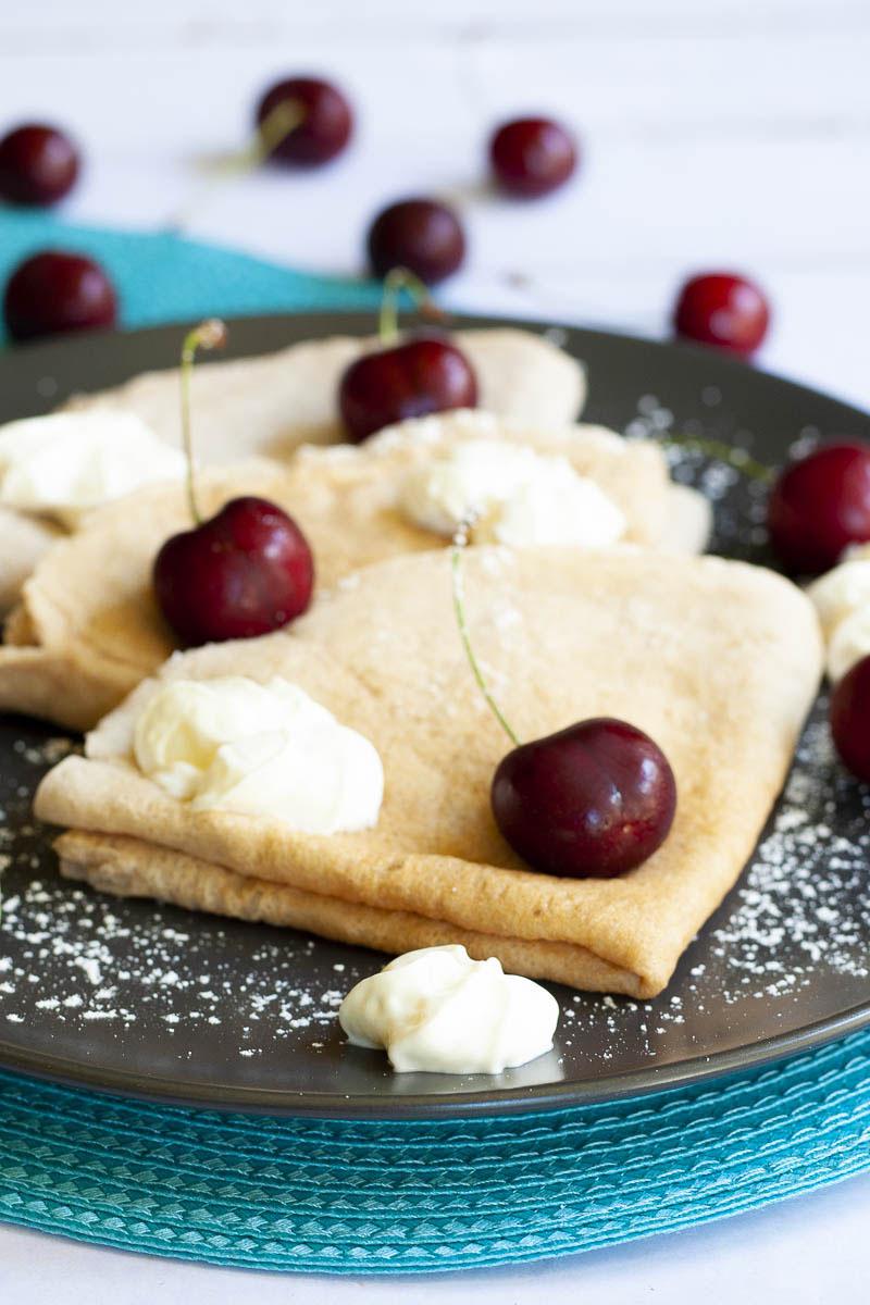 Black plate from top with 3 crepes folded as triangles with cherry and whipped cream scattered on top. 