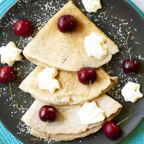 Black plate from top with 3 crepes folded as triangles with cherry and whipped cream scattered on top.