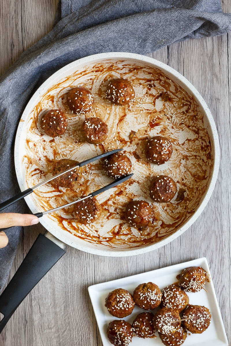 Frying pan from above with several brown-red balls sprinkled with sesame seeds. A hand holding tongs takes one ball.