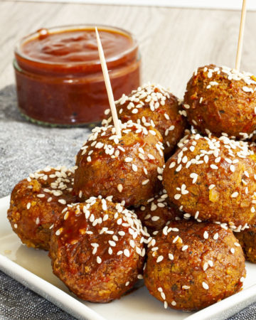 White plate with several brownish-redish balls on top of each other sprinkled with sesame seeds. In the background a small glass jar with red sauce.