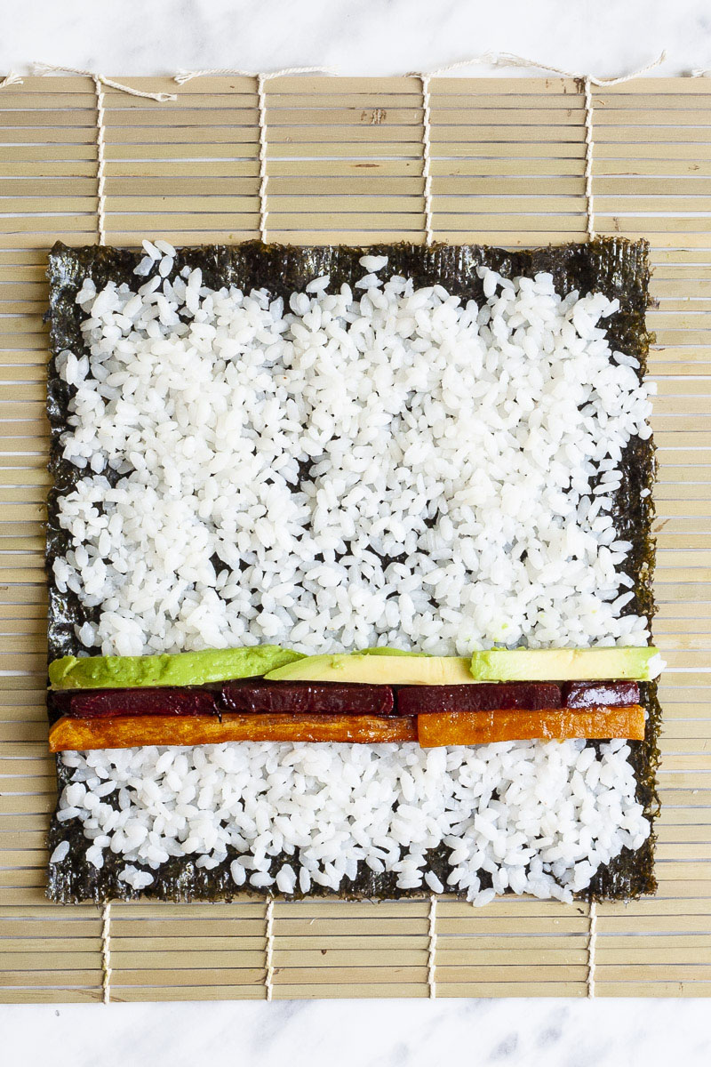  Tapis en bambou avec une feuille en forme de papier vert surmontée de riz blanc et de bâtonnets colorés de légumes verts violets et jaunes formant 3 lignes. 