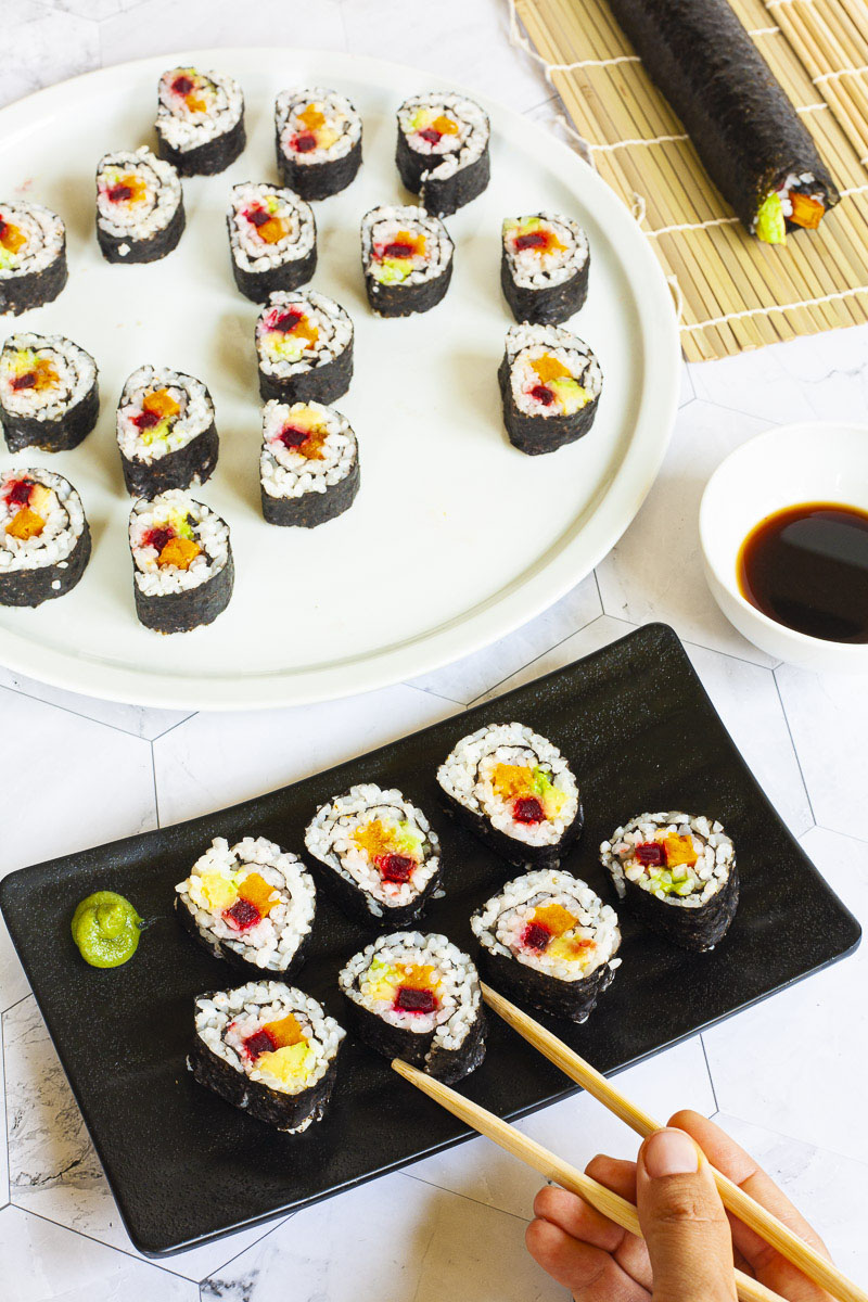 7 sushi rolky na černém talíři. Rohlíky mají černý kryt a bílý uvnitř s fialově žlutou a zelenou uprostřed. Velká bílá deska vzadu s více rolkami. 