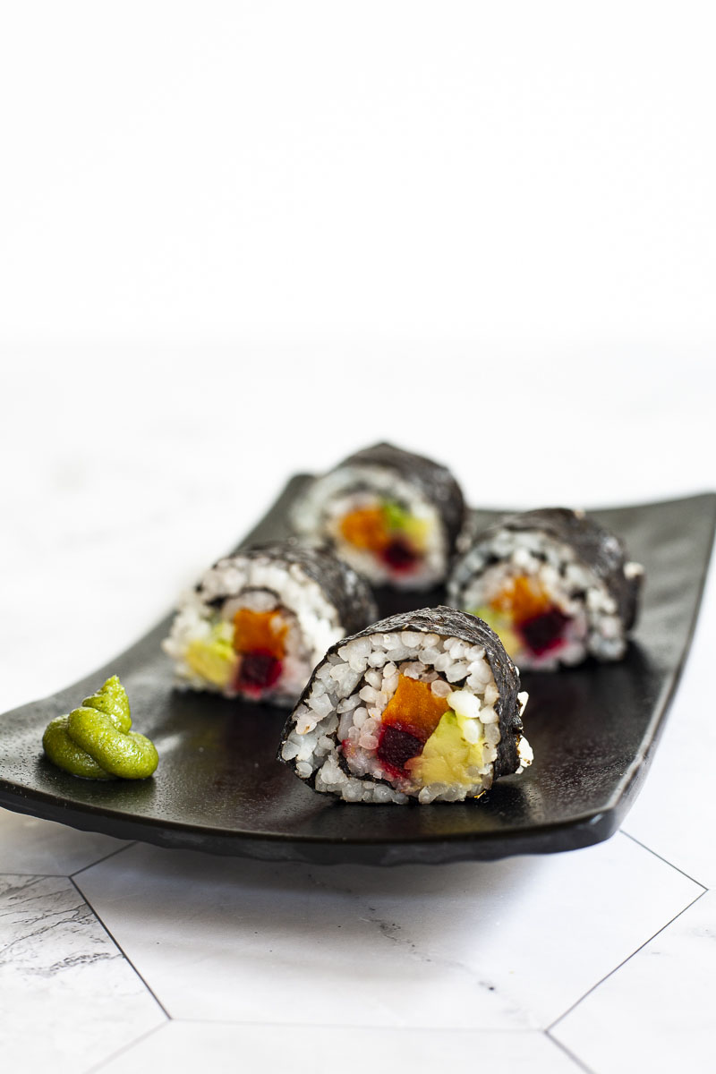 4 sushi broodjes op een zwart bord. De rollen hebben een zwarte cover en een witte binnenkant met paars geel en groen in het midden.