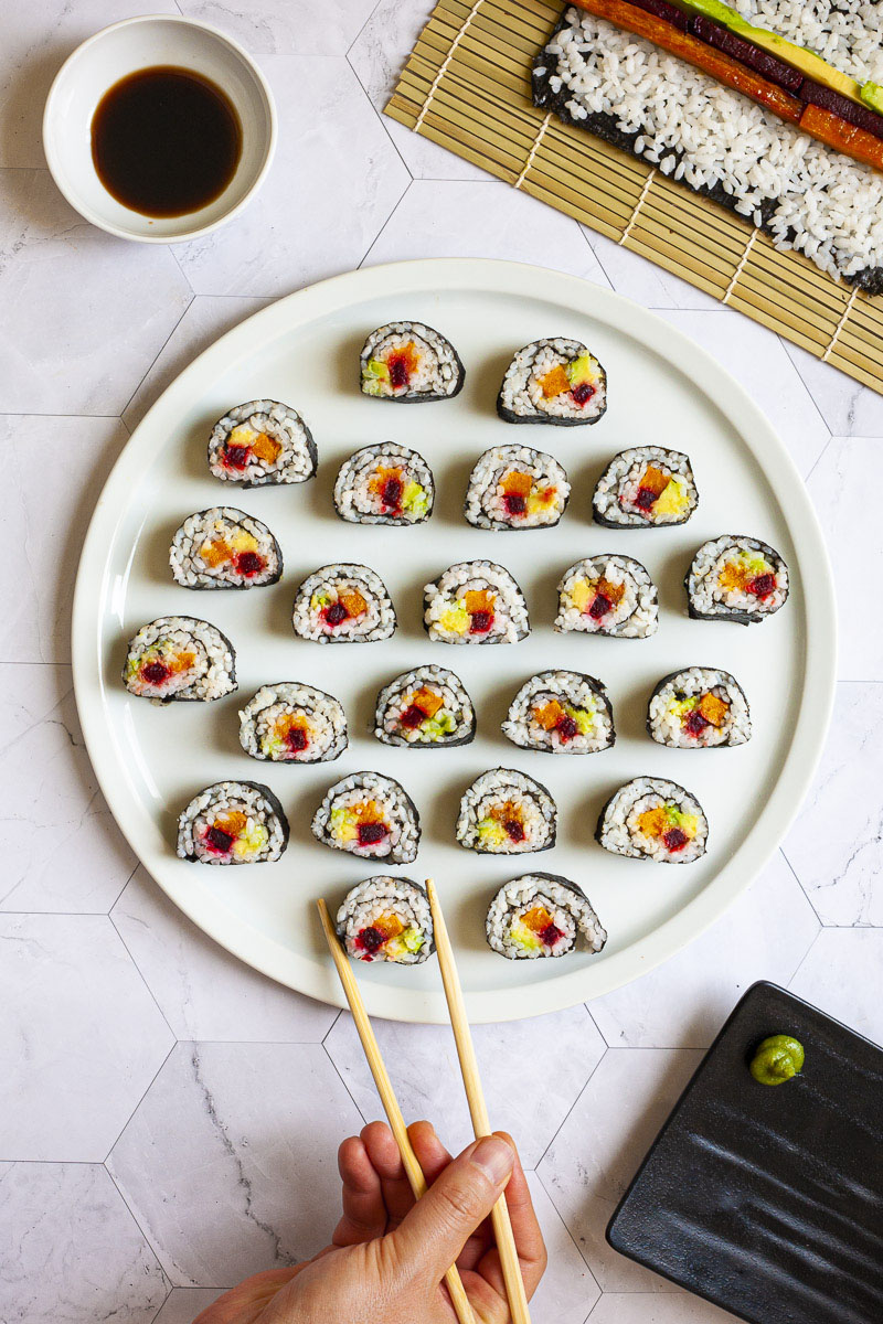 Numerosi rotoli di sushi su un grande piatto bianco rotondo. I rotoli hanno coperchio nero e bianco all'interno con viola giallo e verde nel mezzo.