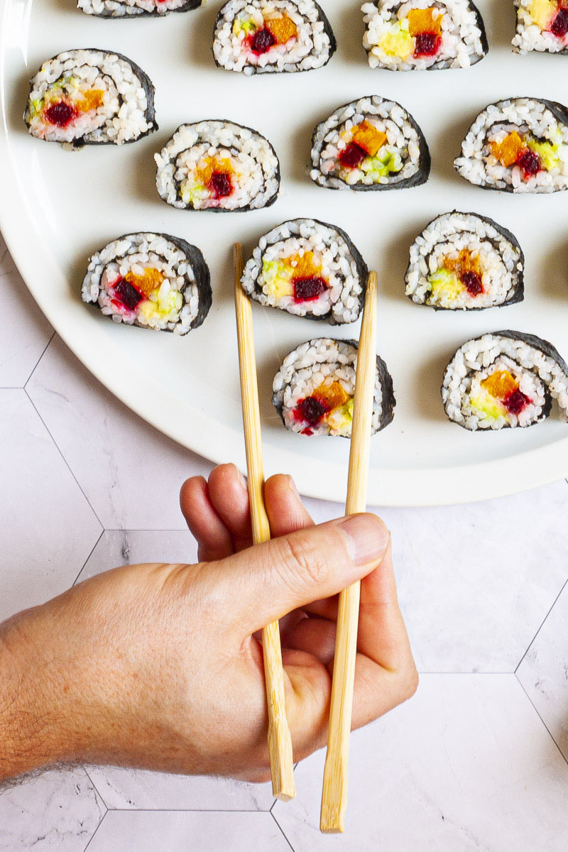 A hand is holding chopsticks and reaching for one sushi on a large white plate. The rolls have black cover and white inside with purple yellow and green in the middle.