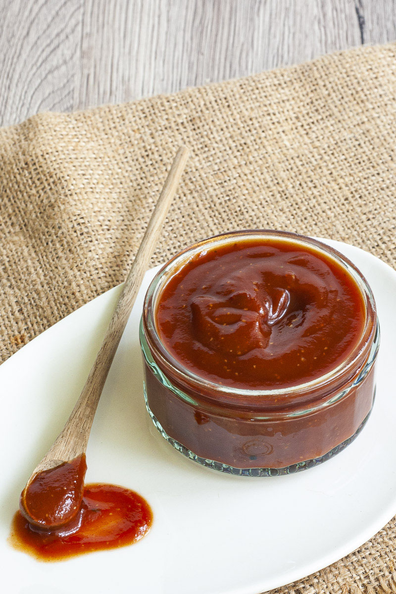 Small glass container full of red thick sauce. A small wooden spoon is laid next to it on a white plate dripping with red sauce. 
