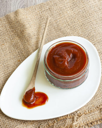 Small glass container from above full of red thick sauce. A small wooden spoon is laid next to it on a white plate dripping with red sauce.