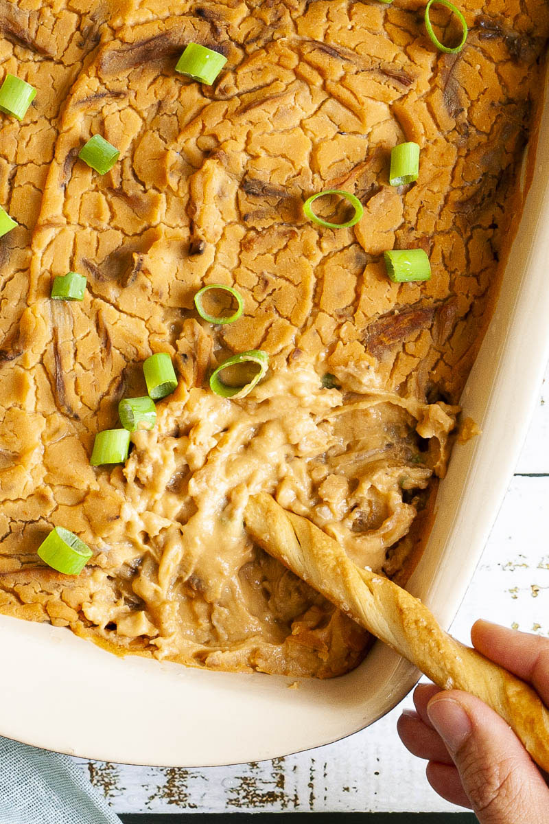 In a light brown square dish there is an orange dip with brown shreds and green spring onion rings. A hand is dipping a breadstick in the corner. 