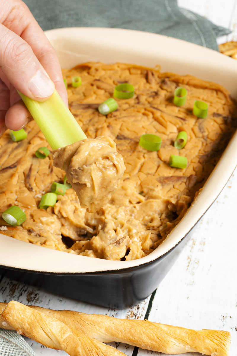 A hand is holding a green celery stick that was dipped in an orange dip with brown shreds. The dip is at the background in a light brown square dish.