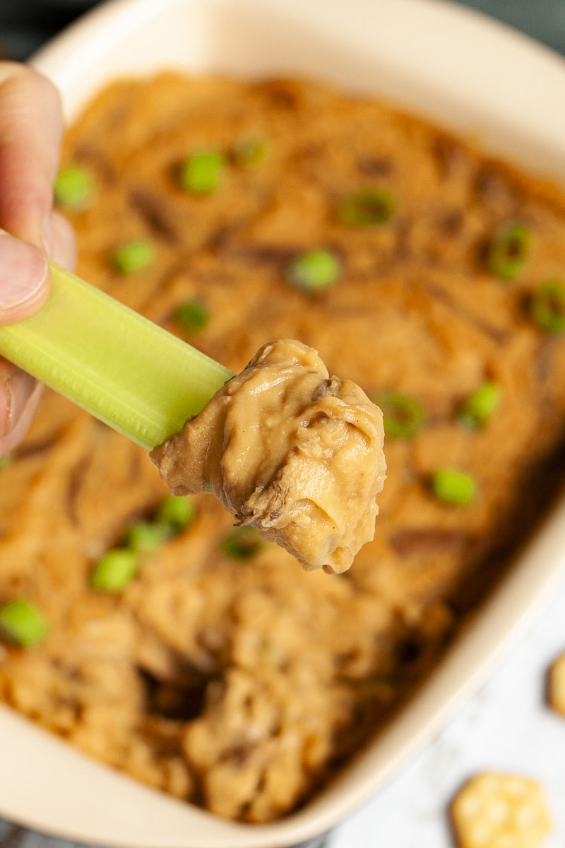 A hand is holding a green celery stick that was dipped in an orange dip with brown shreds. The dip is at the background in a light brown square dish.