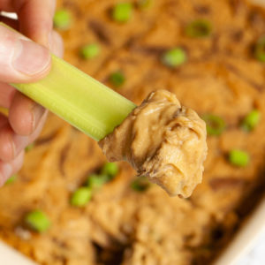 A hand is holding a green celery stick that was dipped in an orange dip with brown shreds. The dip is at the background in a light brown square dish.