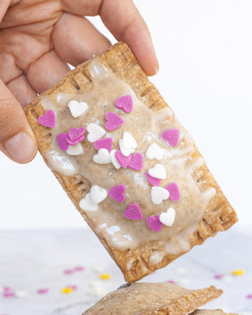 A hand is holding a pop tart, a rectangle-shaped pillow-like cookie with white glaze on top and pink and white small hearts