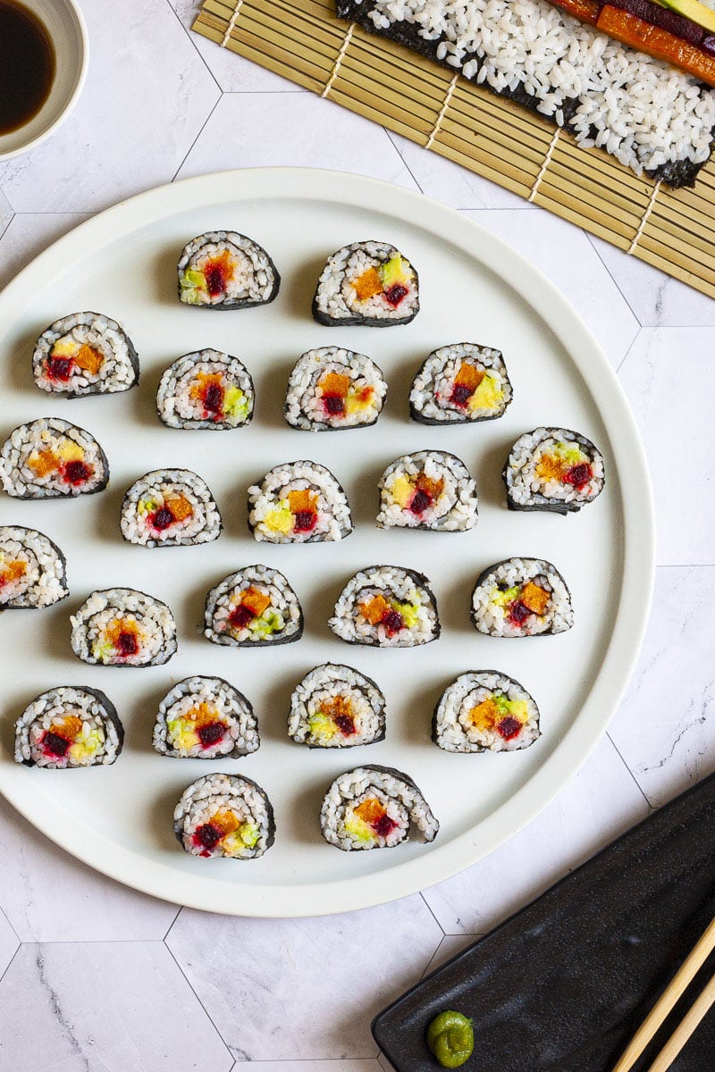 Numerous sushi rolls on a large round white plate. The rolls have black cover and white inside with purple yellow and green in the middle.