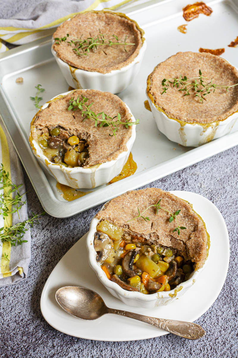 4 small white pots covered with brown pie crust and topped with green herbs. 2 of them has some crust missing and you can see the veggie stew within the yellow, green, orange, brown veggies of corn, green peas, mushrooms and carrots