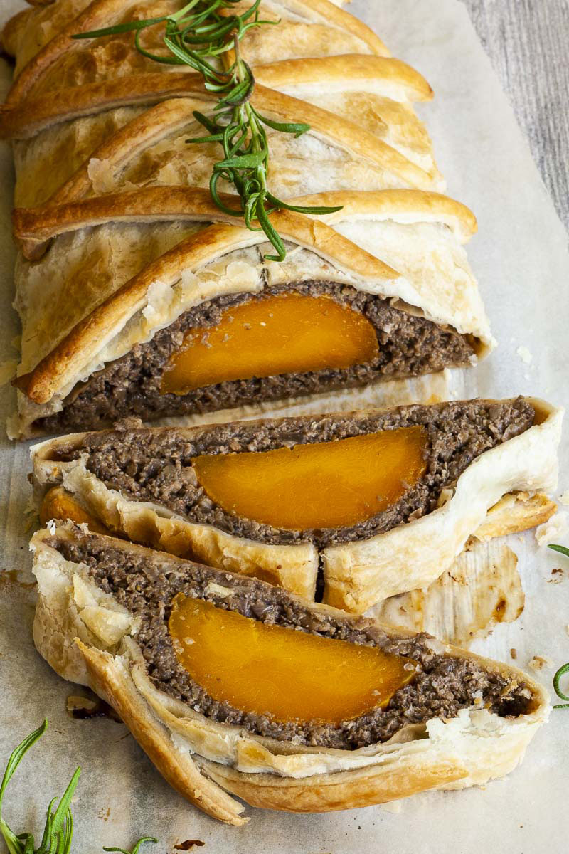 2 slices of vegan wellington on the side. Yellow squash with brown puree around wrapped in a pastry shell. Green herbs are around.