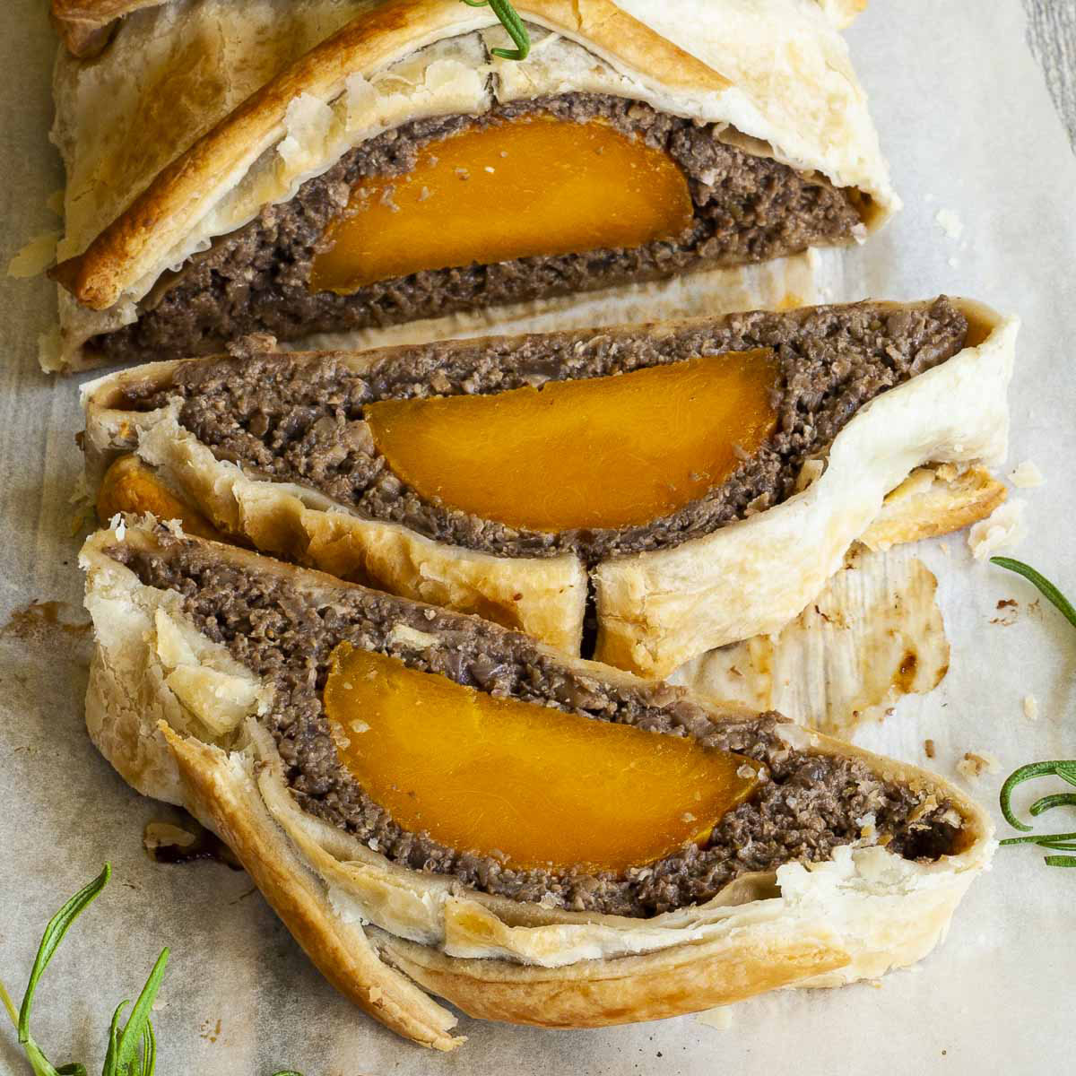 2 slices of vegan wellington on the side. Yellow squash with brown puree around wrapped in a pastry shell. Green herbs are around.