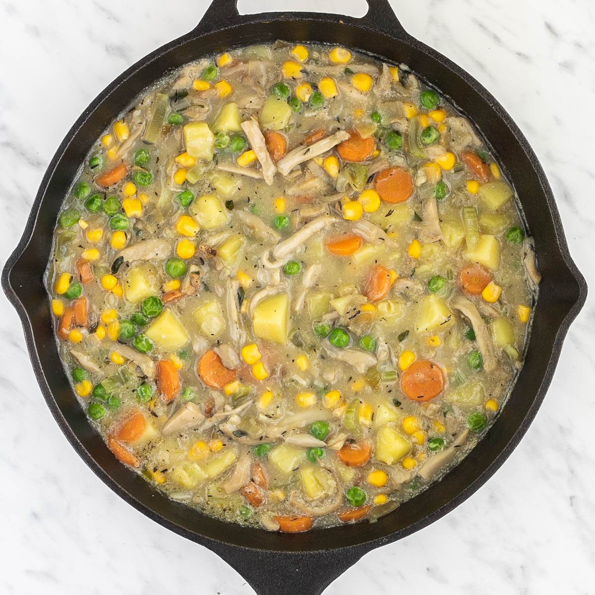 Cast iron skillet with light brown thick stew full of sliced carrots, chopped onion, garlic, celery, potatoes, corn, peas, shredded king oyster mushrooms, bay leaves, thyme.