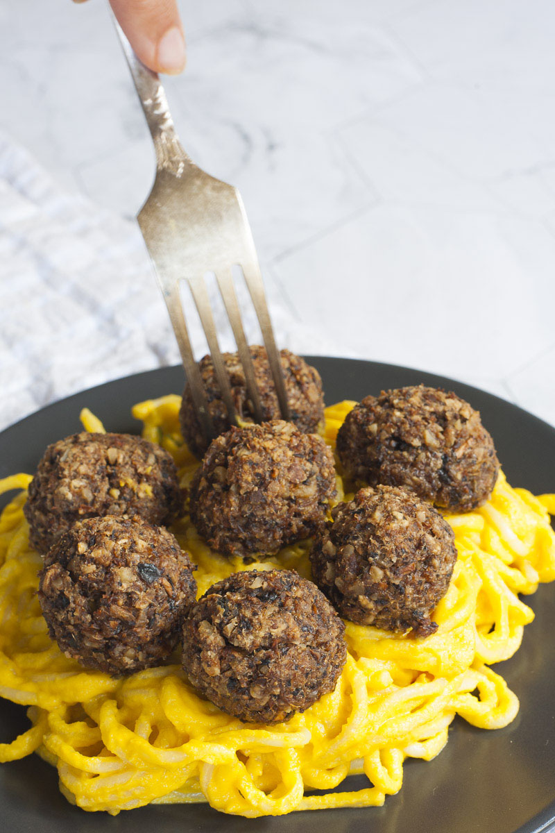 Several mushroom meatballs on top of spaghetti with yellow creamy sauce served on a black plate. A fork is taking on meatballs from the middle.