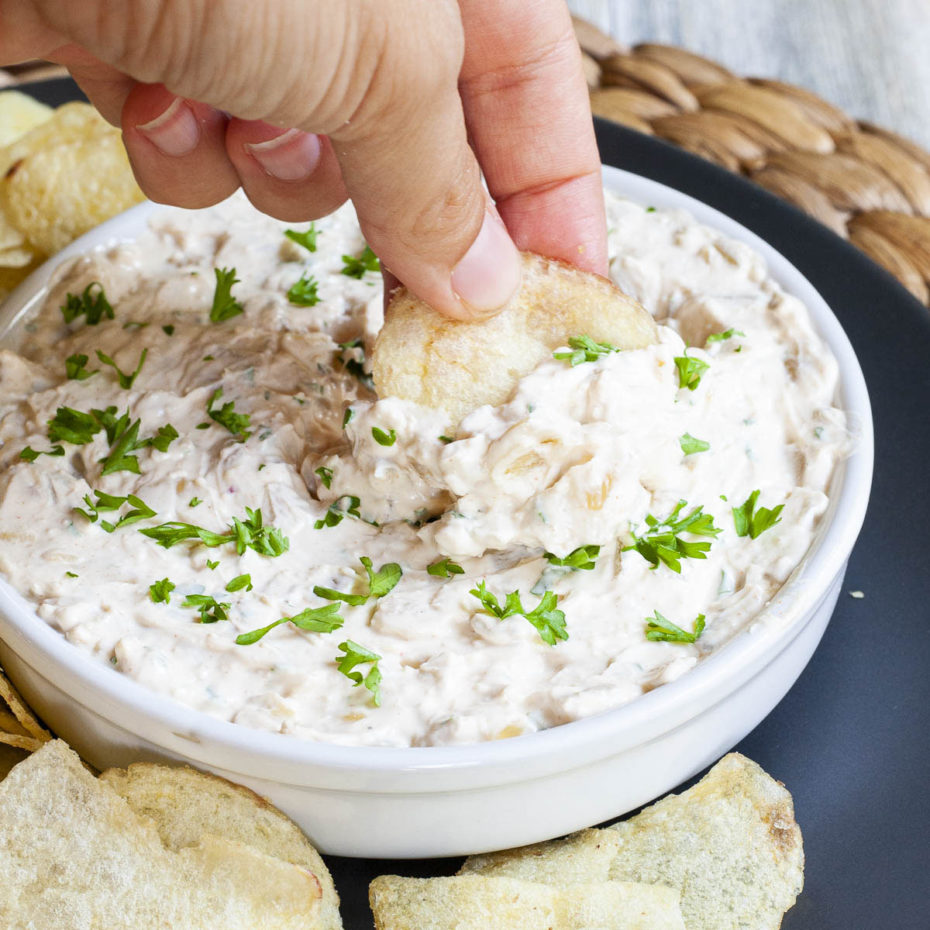 White bowl with white thick creamy sauce in it sprinkled with fresh green herbs. A hand digs a potato chips in the middle.