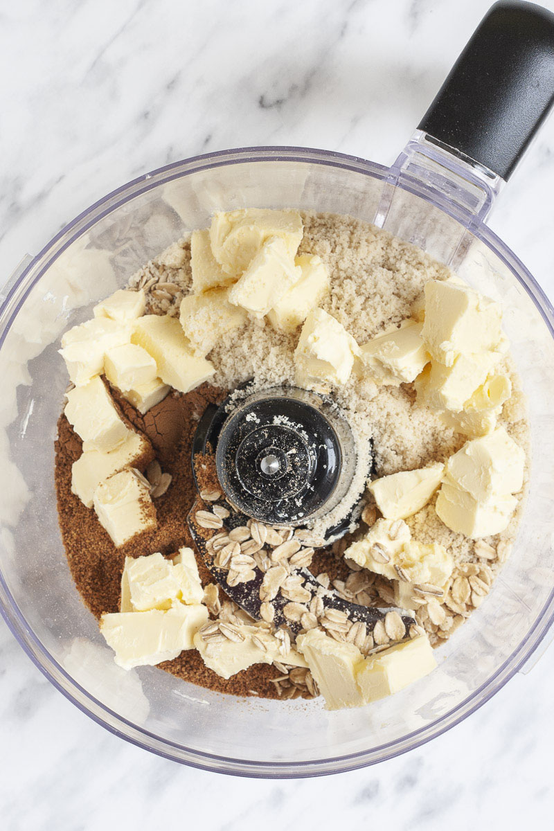 A food processor from above showing the blades, brown sugar, rolled oats, diced butter and light brown flour.