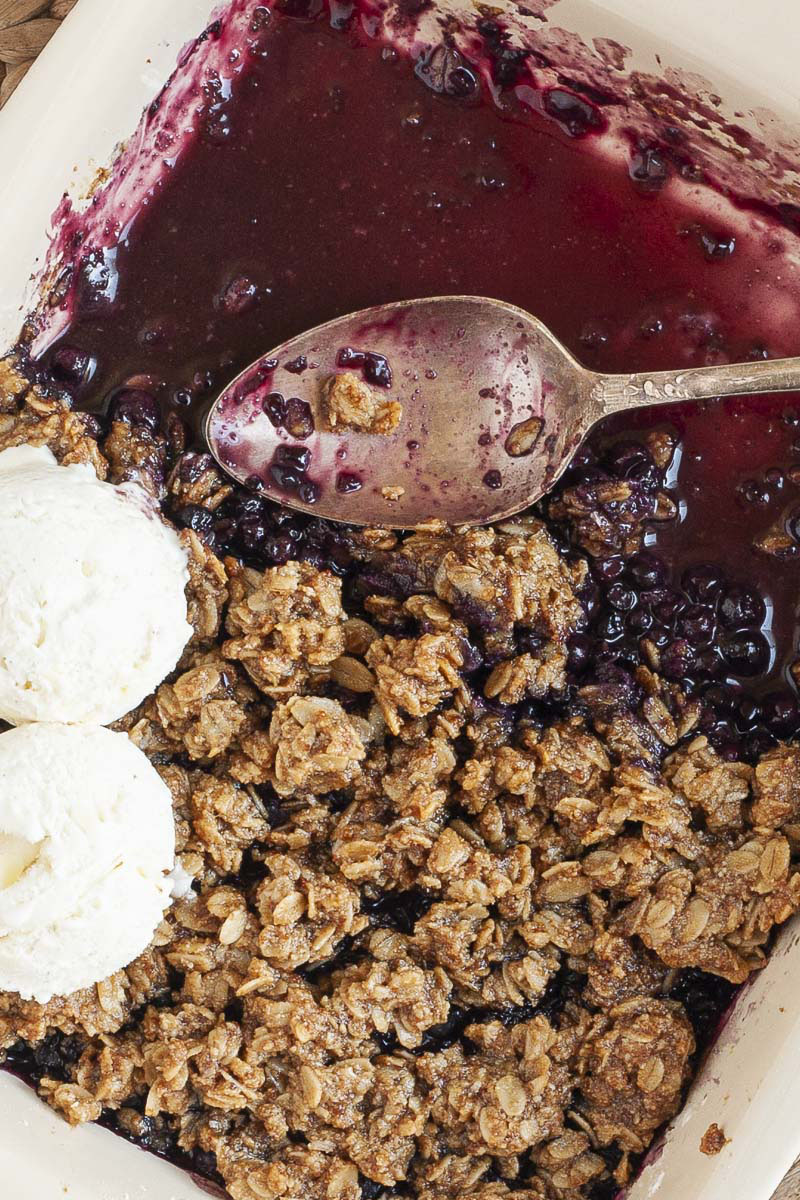 A spoon is in the middle of a white rectangle baking dish. Half of the dish is full of brown crisp, the other half is empty only the purple juices of the blueberry colors the bottom of the dish.