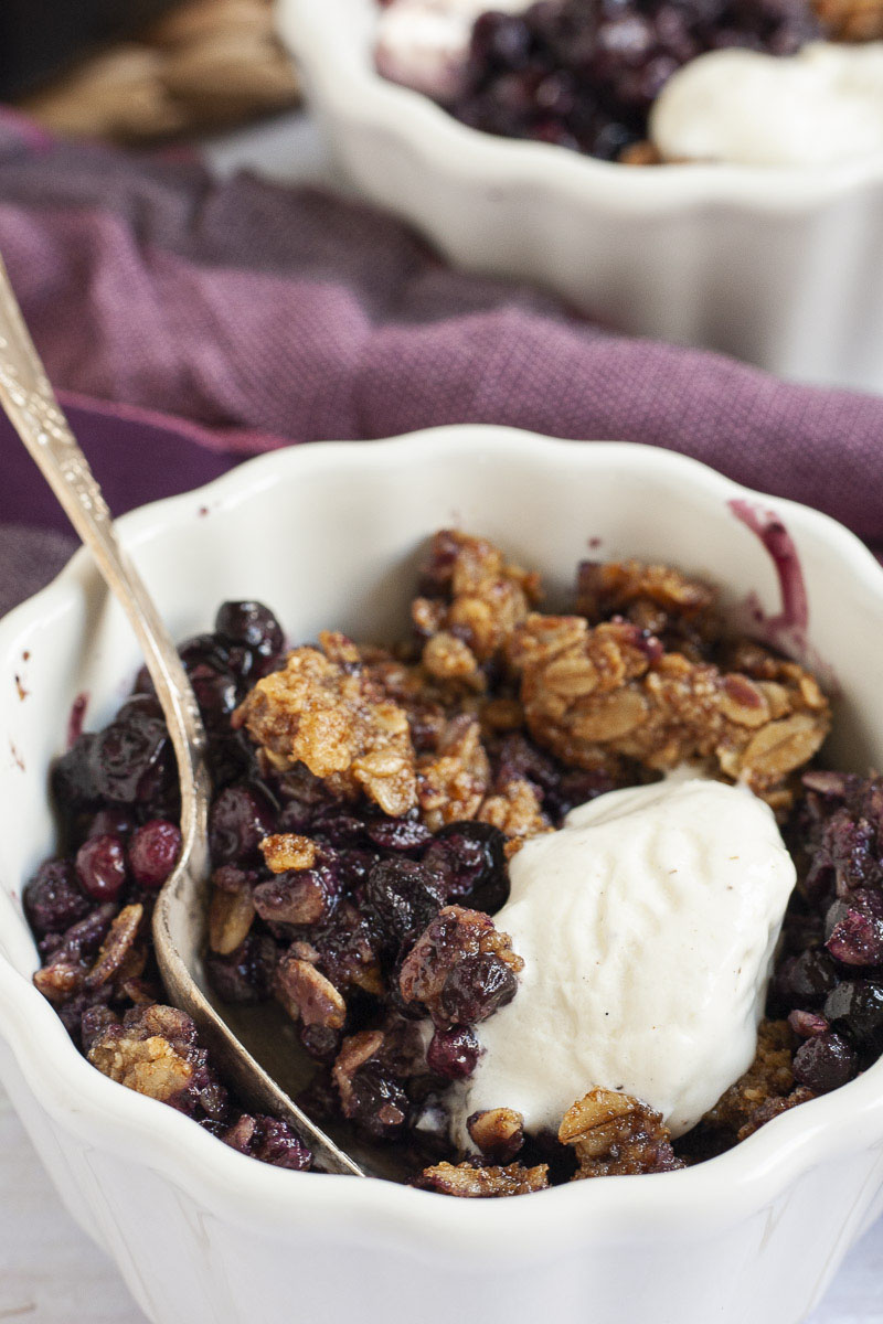 Up close a small white bowl with one serving of purple blueberries, golden brown crisps and a small scoop of white ice cream.