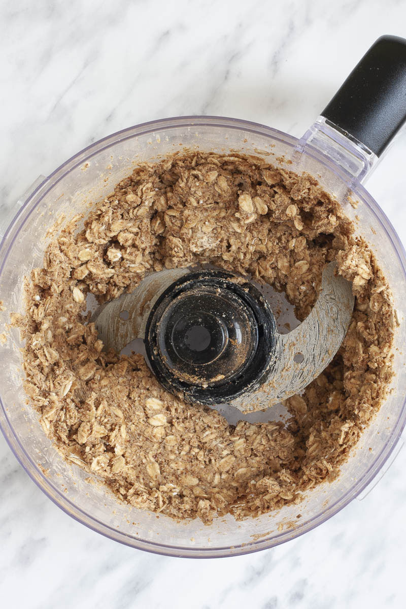 A food processor from above showing the blades and a brown mixture with rolled oats in it.