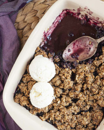 A spoon is in the middle of a white rectangle baking dish. Half of the dish is full of brown crisp, the other half is empty only the purple juices of the blueberry colors the bottom of the dish.