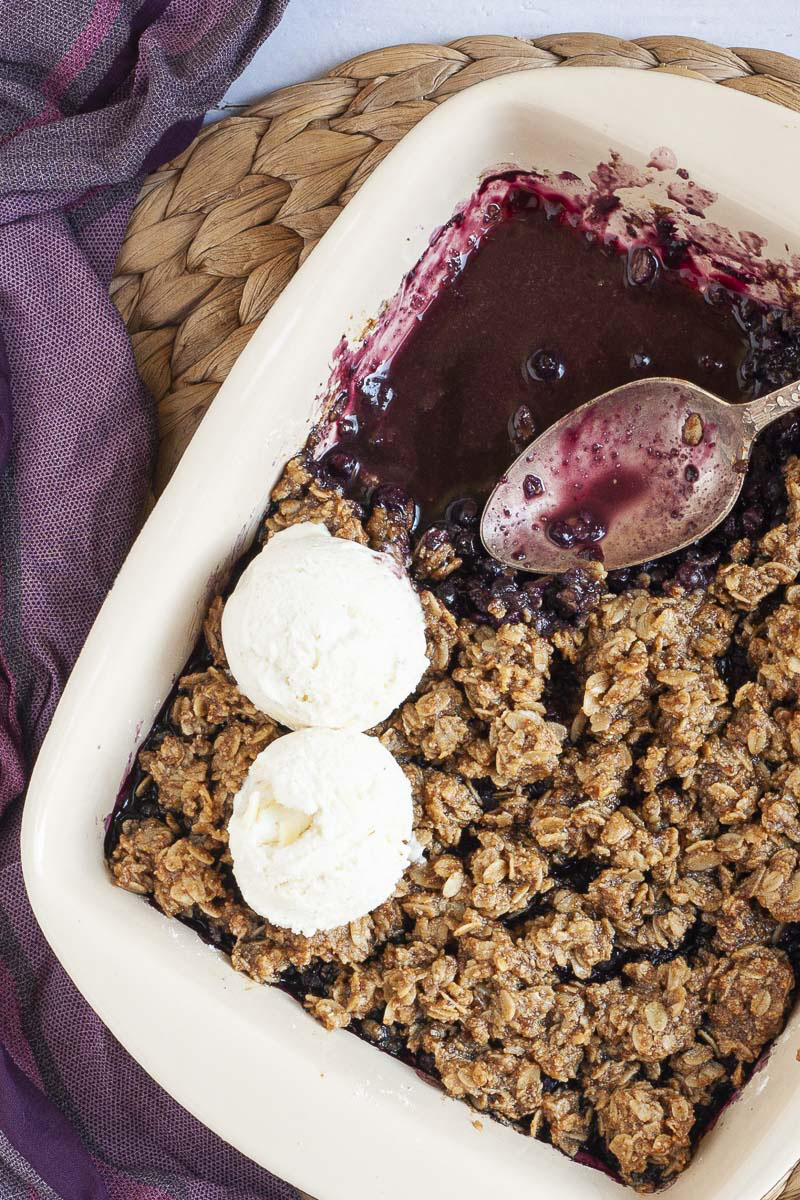 A spoon is in the middle of a white rectangle baking dish. Half of the dish is full of brown crisp, the other half is empty only the purple juices of the blueberry colors the bottom of the dish.