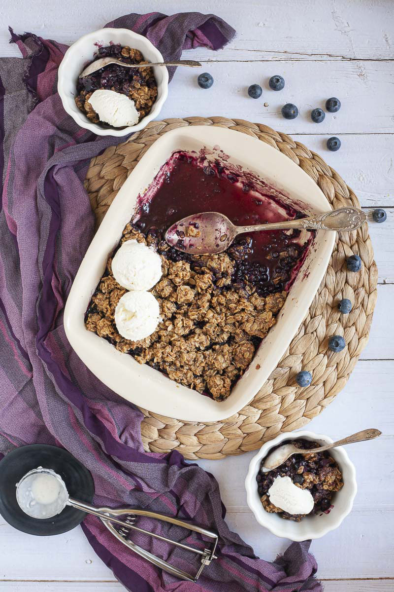 From above spoon is in the middle of a white rectangle baking dish. Half of the dish is full of brown crisp, the other half is empty only the purple juices of the blueberry colors the bottom of the dish. 2 small bowls with 1 serving each is around it. Scattered blueberries everywhere