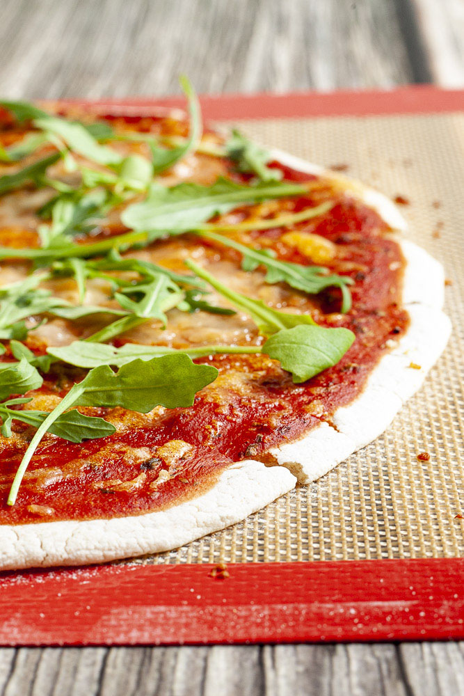 Gluten-free pizza with tomato sauce, melted cheese and fresh arugula on a red brown baking sheet