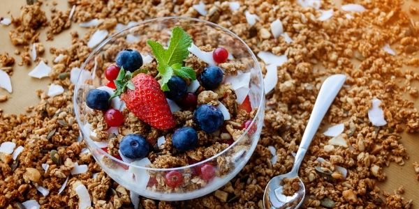 A small glass bowl with yoghurt, granola, and berries. Granola scattered around