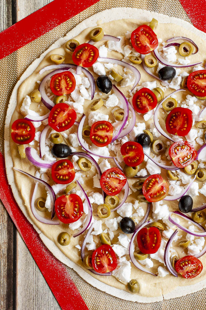 Large thin pizza crust rolled out on top of a red brown silicone rolling mat topped with hummus, cherry tomatoes, purple onion slices, green and black olives and feta cheese crumble before baking