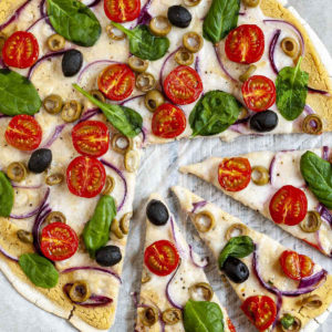 Baked and sliced pizza from above on white parchment paper topped with hummus, cherry tomatoes, purple onion slices, green and black olives, melted cheese and fresh basil leaves.
