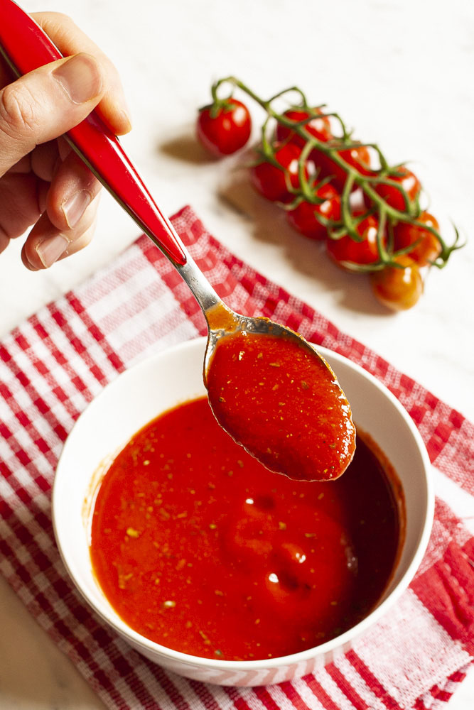 Red pizza sauce in a white bowl. A hand is holding a spoon taking some of the sauce closer. Red cherry tomato vine is in the background