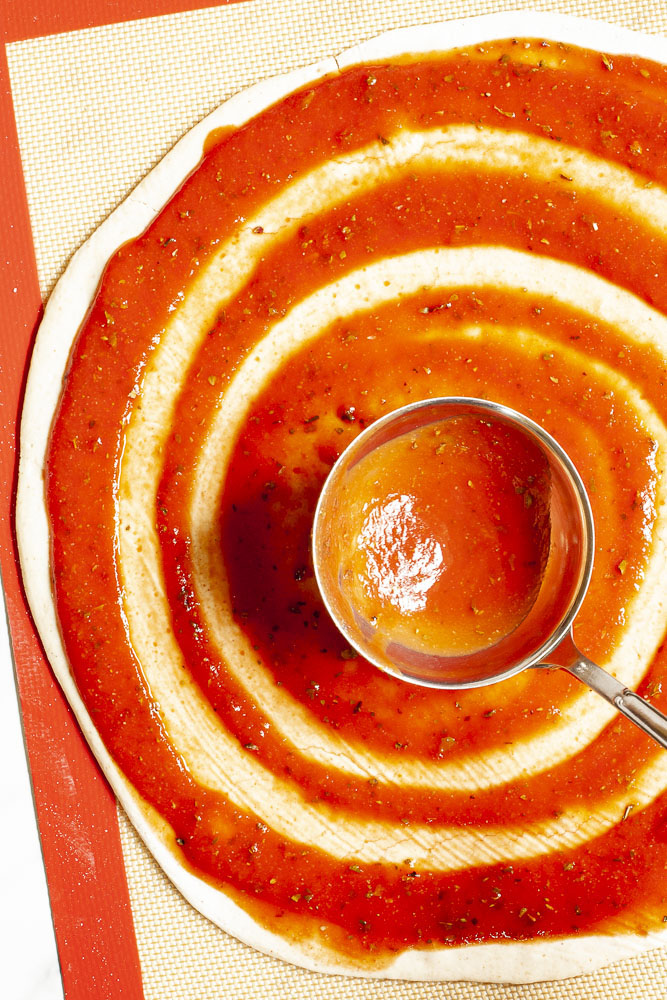 Red pizza sauce is spread in circles on top of a pizza crust with a ladle.