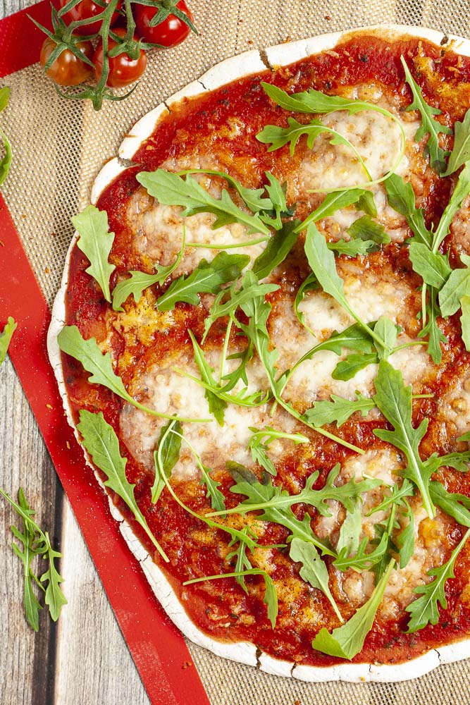 Gluten-free pizza with tomato sauce, melted cheese and fresh arugula on a red brown baking sheet from above.