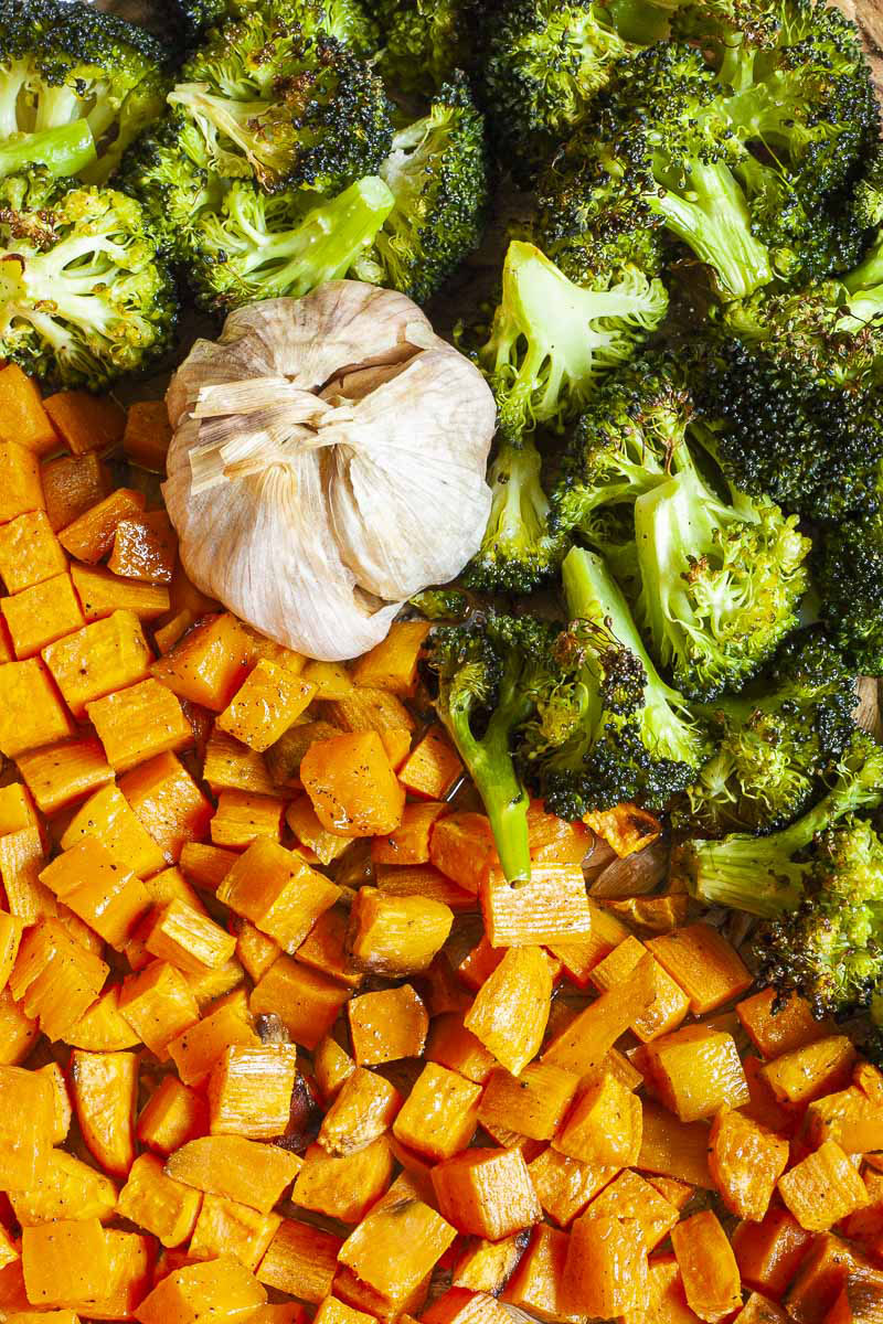 Roasted broccoli florets and dices sweet potato with a whole garlic bulb up really close.